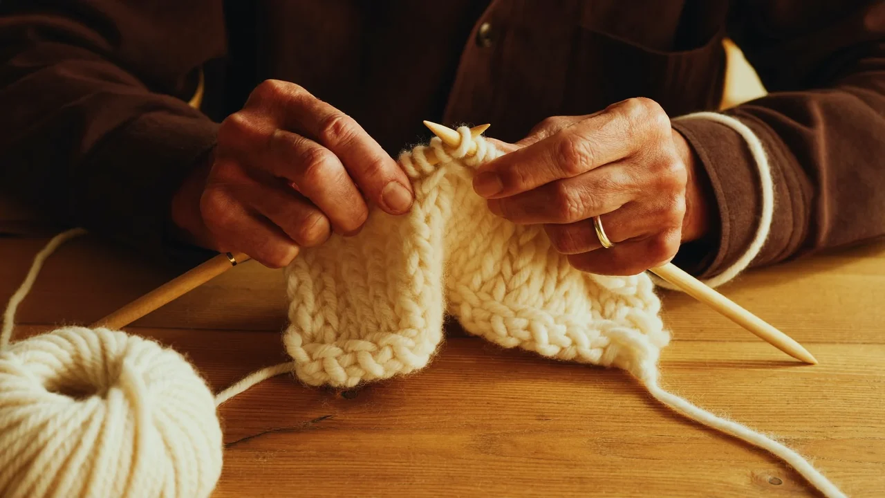 Hände beim Stricken mit cremefarbener Wolle und Holznadeln.